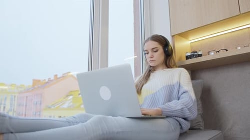 Young woman sitting by the window at home in headphones works at a laptop. Remote study. Remote work