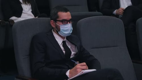 Man in Medical Mask Taking Notes in Notebook at Conference