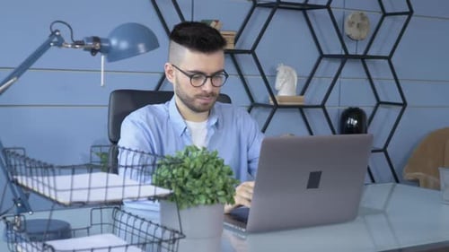 Young handsome man in glasses working on his laptop and sitting in the office at home