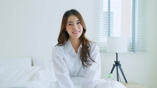 Young Woman Smiles, Sitting in Bed in White Shirt