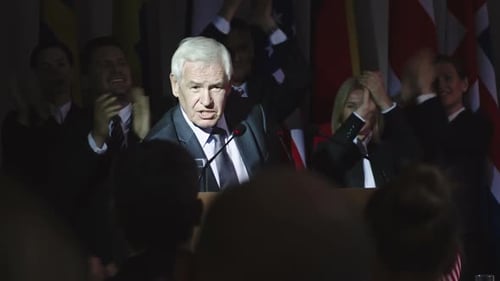 Politician Speaks to Audience at Podium with Flags