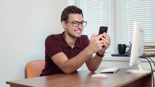 Man Looking at the Screen of His Phone and Smiling Texting in the Office Sitting Table