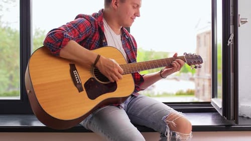 Young Man Playing Guitar Sitting on Windowsill