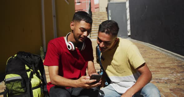 Two mixed race friends sitting and using smartphone in the street ...