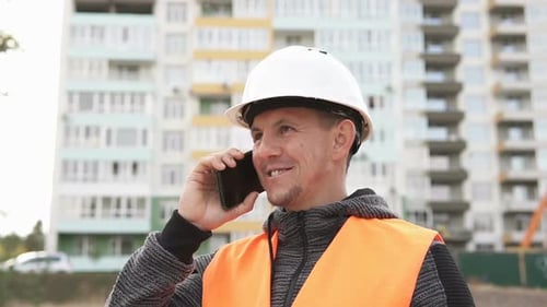 The foreman at the construction site is talking on the phone and checking the drawings.