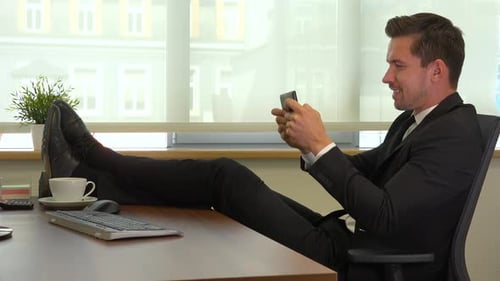 An office worker sits in front of a computer with legs on the desk and plays a game on smartphone