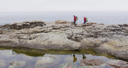 Senior hiker couple with backpacks walking on the rocks while hiking near sea shore.