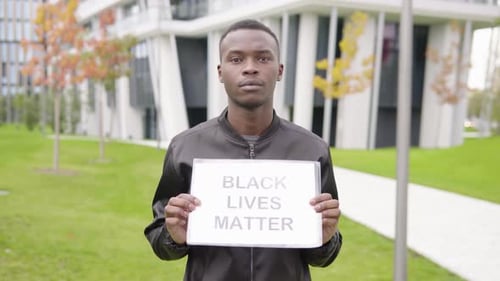 A Young Black Man Shows a Black Lives Matter Sign To the Camera - Office Buildings