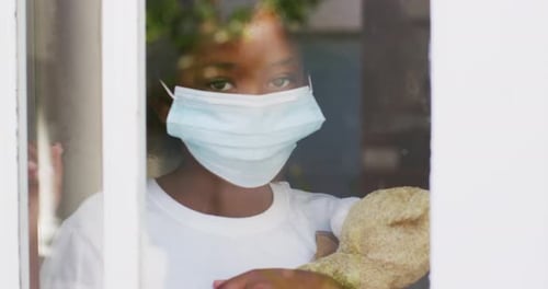 Child Wearing Mask Holding Toy Behind Window
