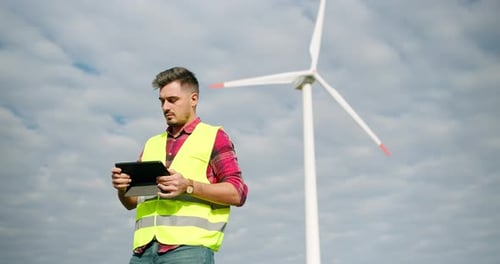The Engineer Work Next to the Windmill Turbines with the Tablet in His Hand