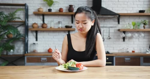 Asiatische Frau sitzt am Tisch und sieht traurig und gelangweilt mit Diät aus und will keinen Salat essen