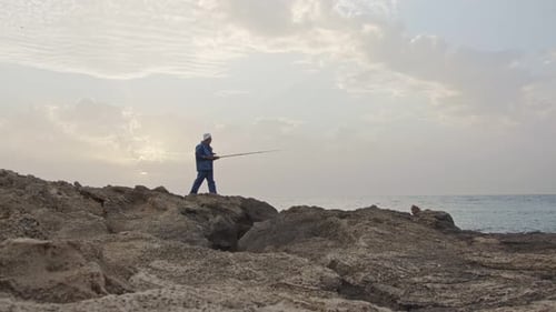 Old fisherman standing on sea side rocks and fishing against the sunset