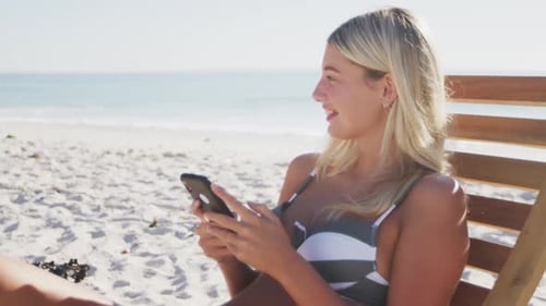 Caucasian woman sitting on a sunbed and using her smartphone on the beach