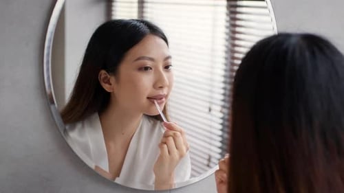 Young Woman Applying Lip Gloss in Bathroom Mirror