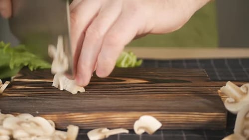 Die männlichen Hände des Kochs schneiden frische Champignons auf einem braunen Holzbrett mit Hilfe eines