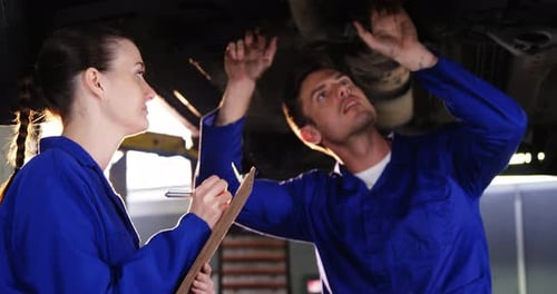 Female mechanic writing on clipboard while male mechanic checking a car