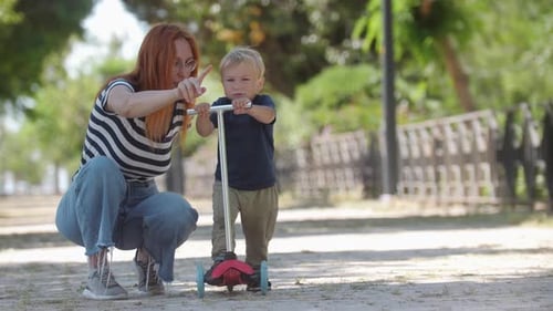 Woman Teaching Child To Ride Scooter Outdoors