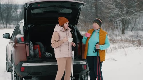 Women Talk and Drink Tea From a Thermos While Standing Near a Car in a Snowy Area