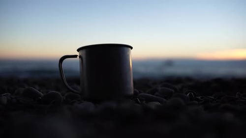 Closeup of a Travel Stainless Mug Standing on a Rock Against a Backdrop of a Beach and Calm Sunset