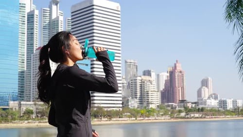 4K Asian woman drinking water from a bottle while jogging at public park in the morning.