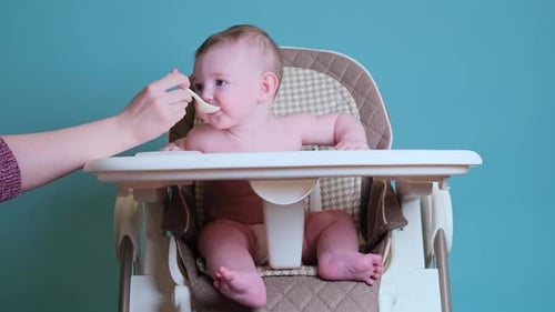 Mother feeding toddler baby with spoon on high chair for children, blue studio background