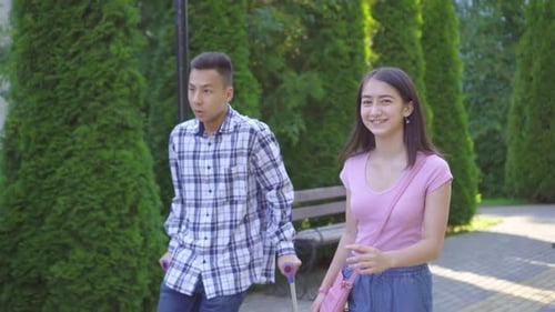 Young Asian Man with Broken Leg on Crutches Communicates with Young Asian Woman in Park