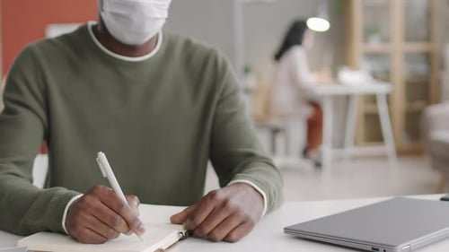 Adult Wearing Mask Writes in Notebook at Desk