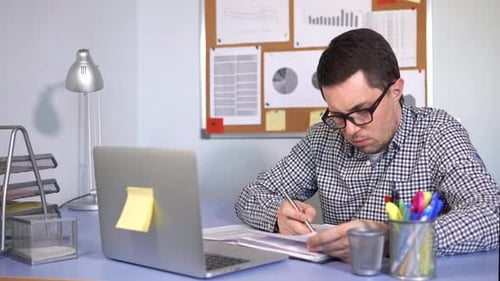 Office Worker Working on Laptop and Writing Down Information in Papers.