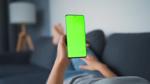 Woman at Home Lying on a Sofa and Using Smartphone with Green Mock-up Screen in Vertical Mode. Girl