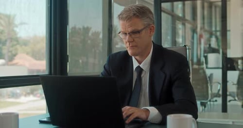 A businessman in formal wear and glasses working on a laptop at the office with big windows.