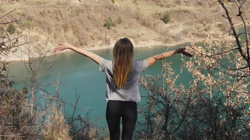 Young Woman Stands with Raised Arms Up on the Nature Lake Sunset Background