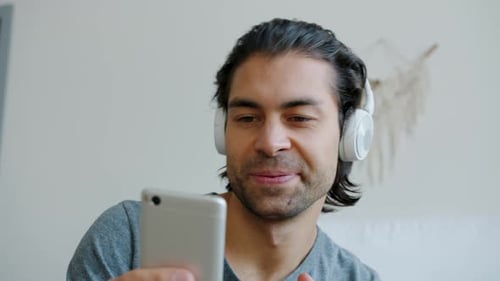 Close-up of Happy Young Brunet Wearing Headphones Enjoying Music and Smartphone Content in Bedroom