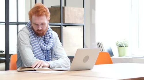 Man Reads Book Next to Laptop at Desk