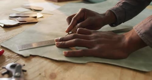 Close up of hands of african american craftsman cutting leather precisely in leather workshop