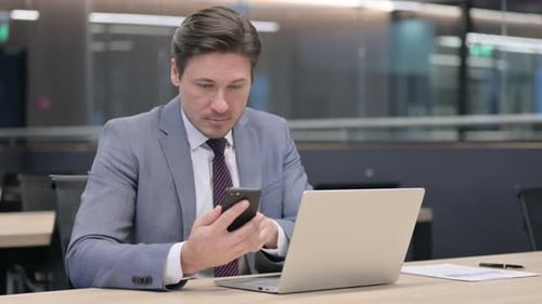 Middle Aged Businessman with Laptop using Smartphone in Office
