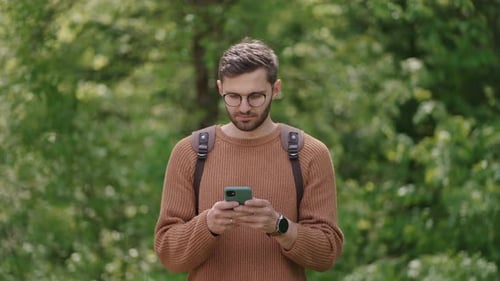 A Man with a Backpack in the Woods on a Mobile Phone is Looking for Information Online Reading the