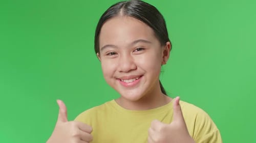 Fröhliches junges asiatisches Mädchen zeigt eine Daumen-Hoch-Geste, während es im Greenscreen-Studio steht