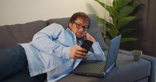 Man Lies on Sofa with Phone and Laptop