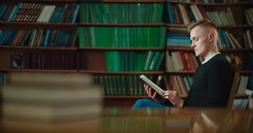 The Guy Is Sitting in a Chair at Library and Slowly Flipping the Book That Interested Him. A