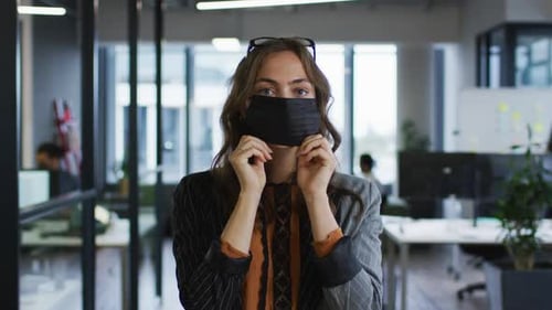 Portrait of caucasian businesswoman adjusting face mask standing in office and looking to camera