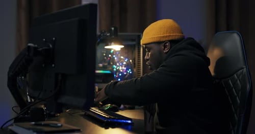 Darkskinned Guy in a Yellow Cap Sits at Desk Getting Ready to Play a Video Game with a Friend Puts