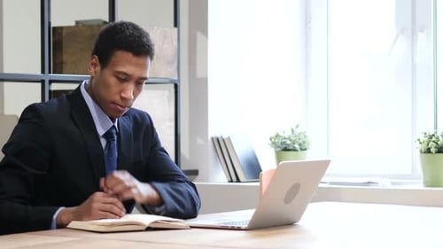 Man Reading Book Next to Laptop in Office