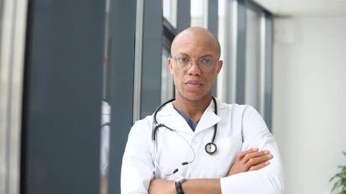 A Young African American Male Doctor Stands in a Medical Suit at the Clinic and Looks at the Camera