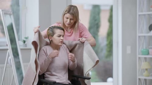 Woman Putting Blanket Over Friend in Wheelchair Indoors