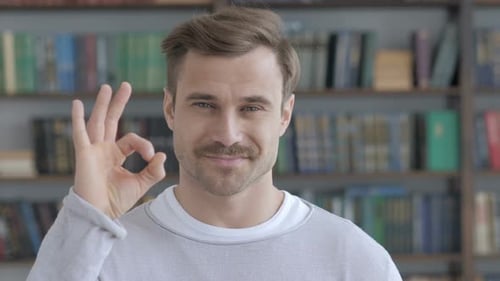 Man Gives Okay Sign in Front of Bookshelf