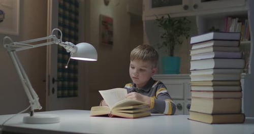 Boy Reads Book Under Desk Lamp at Night