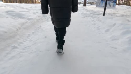 Back View of Woman Legs in Black Clothes and Boots Walking on Winter Snow Path in Park