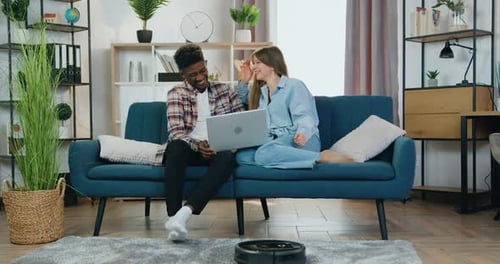 Chico y chica hablando durante la revisión en la computadora de casa mientras un robot aspirador