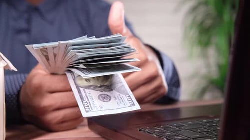 Hand Holding Money Next to House and Laptop