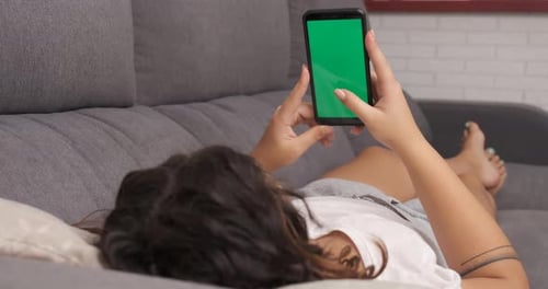 woman on the living room couch watching something on her cell phone, green screen on smart phone